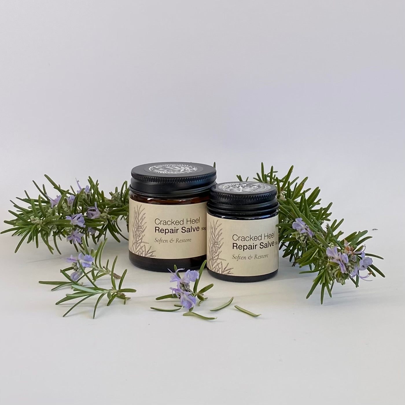 Two jars of Cracked Heel Repair Salve with rosemary leaves and flowers on a white background