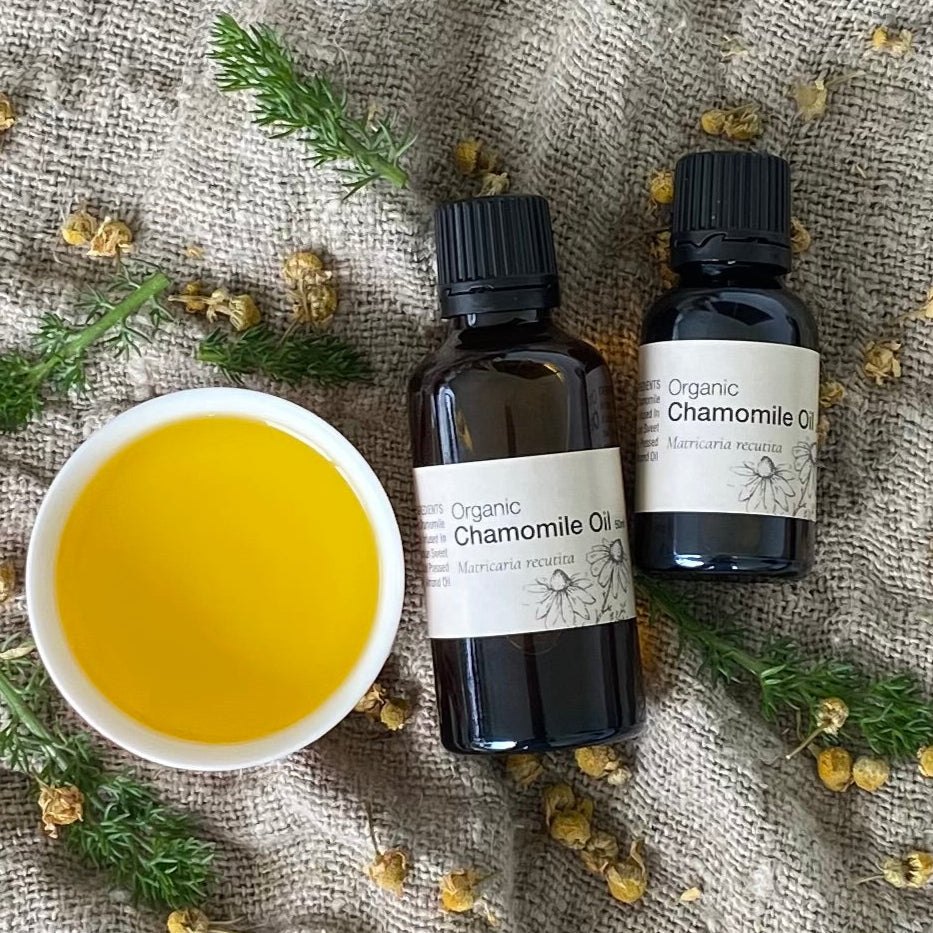 Two bottles of Organic Chamomile Oil with a bowl of oil on a textured fabric background.