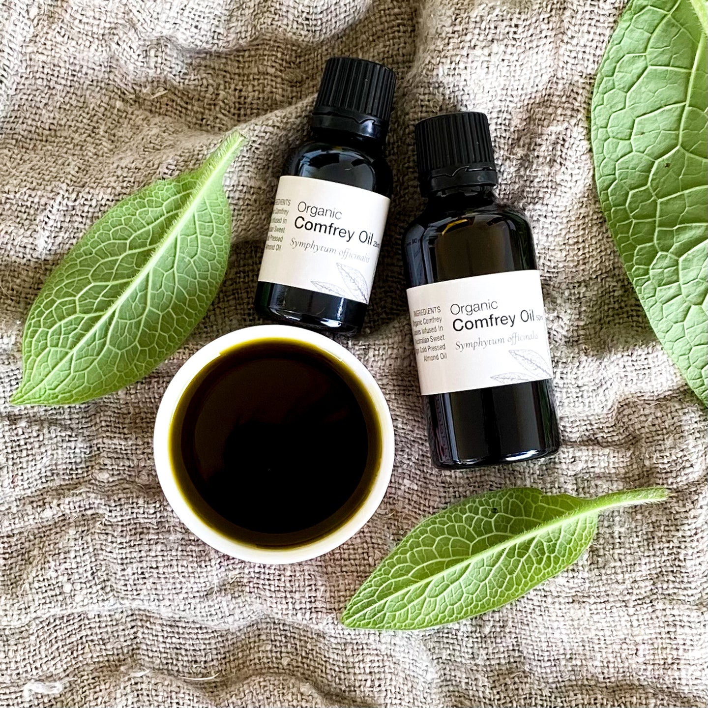 Two bottles of organic comfrey oil with a small bowl of oil and comfrey leaves on a textured fabric background.
