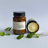 Jar of hydrating antioxidant moisturizer with a lid off, surrounded by green leaves on a light background