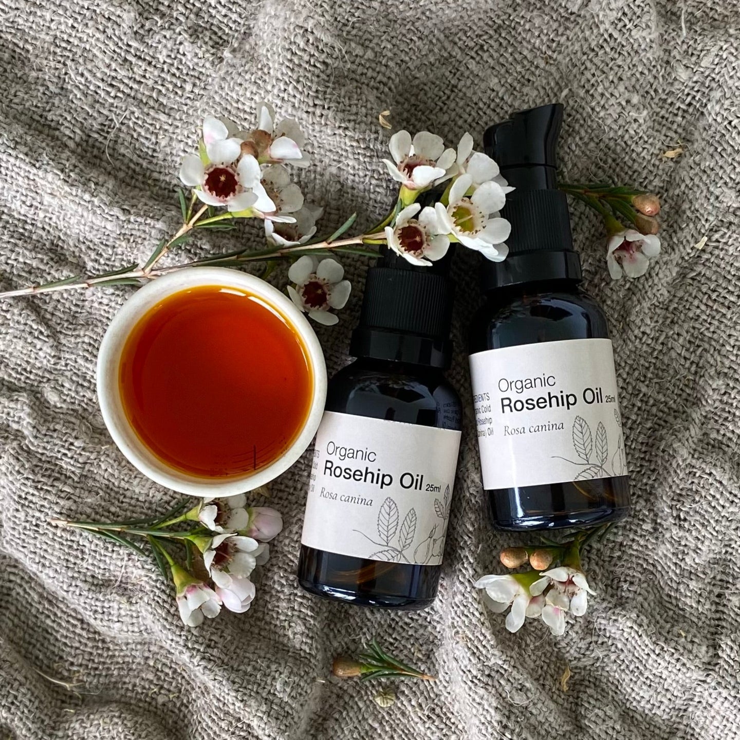 two bottles of organic rosehip oil with a bowl of rosehip oil on a linen background with flowers